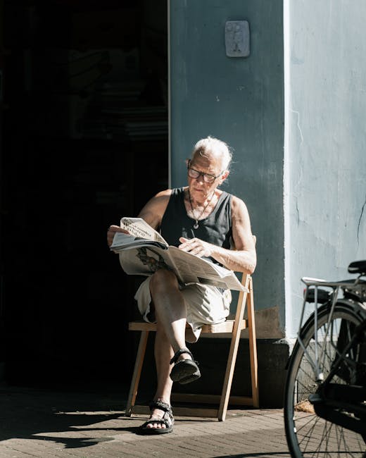 An elderly woman with short grey hair and glasses is sitting outdoors on a wooden chair placed on a concrete pavement, reading a newspaper. She is dressed in a black sleeveless top, beige shorts, and